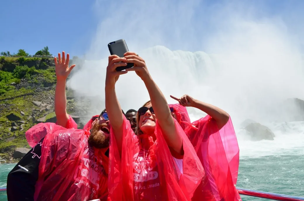 Tourists taking selfie on Niagara Falls boat cruise with waterfall mist and rainbow in background